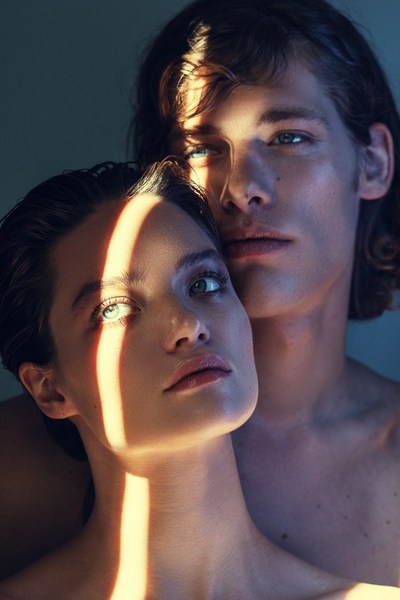 Striking close-up portrait of a young, light-skinned couple with natural makeup, illuminated by a beam of sunlight in a photo studio.