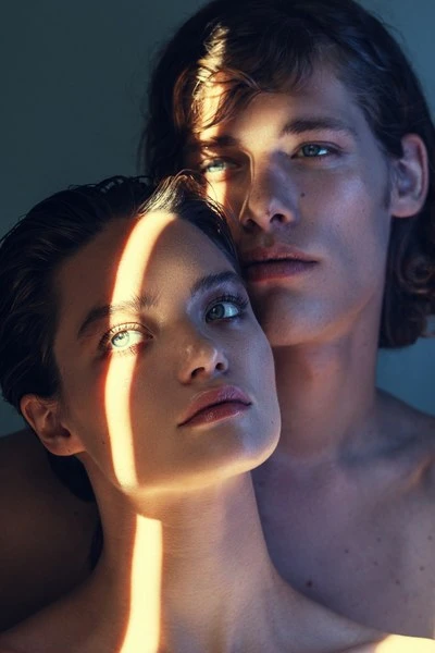 Striking close-up portrait of a young, light-skinned couple with natural makeup, illuminated by a beam of sunlight in a photo studio.