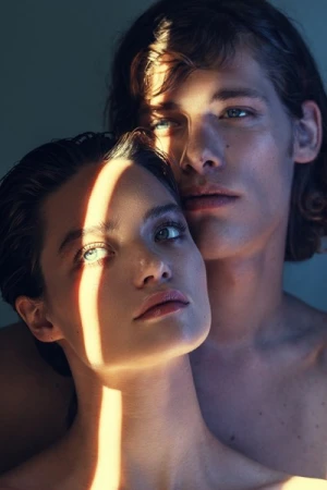 Striking close-up portrait of a young, light-skinned couple with natural makeup, illuminated by a beam of sunlight in a photo studio.