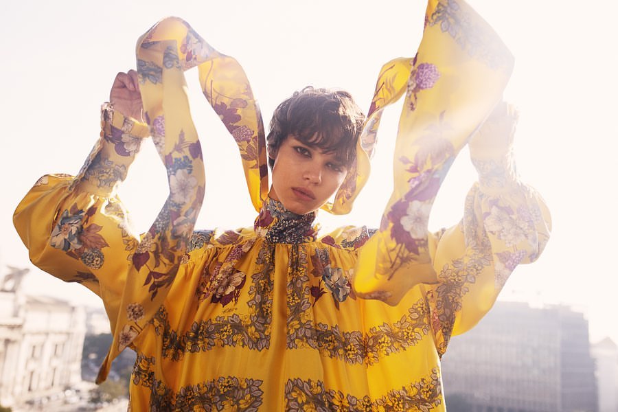 A woman in a bright yellow floral dress poses with hands raised, set against an urban background with visible buildings.