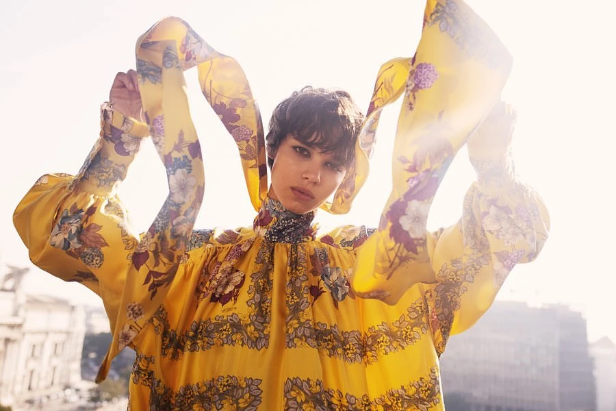 A woman in a bright yellow floral dress poses with hands raised, set against an urban background with visible buildings.