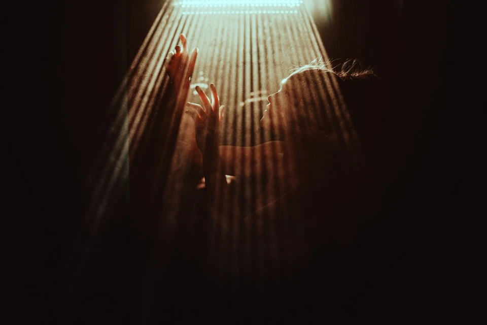 Woman bathed in light, hands raised in a dark setting. Light rays illuminate her silhouette and suggest a sense of mystery and depth.