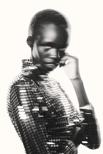 Monochrome portrait of a young woman wearing a square sequined top, with hand near her face, set against a light background.