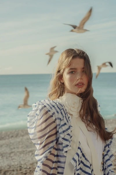 A young woman in a blue and white striped dress stands on a beach with seagulls flying in the background, creating a serene fashion portrait.