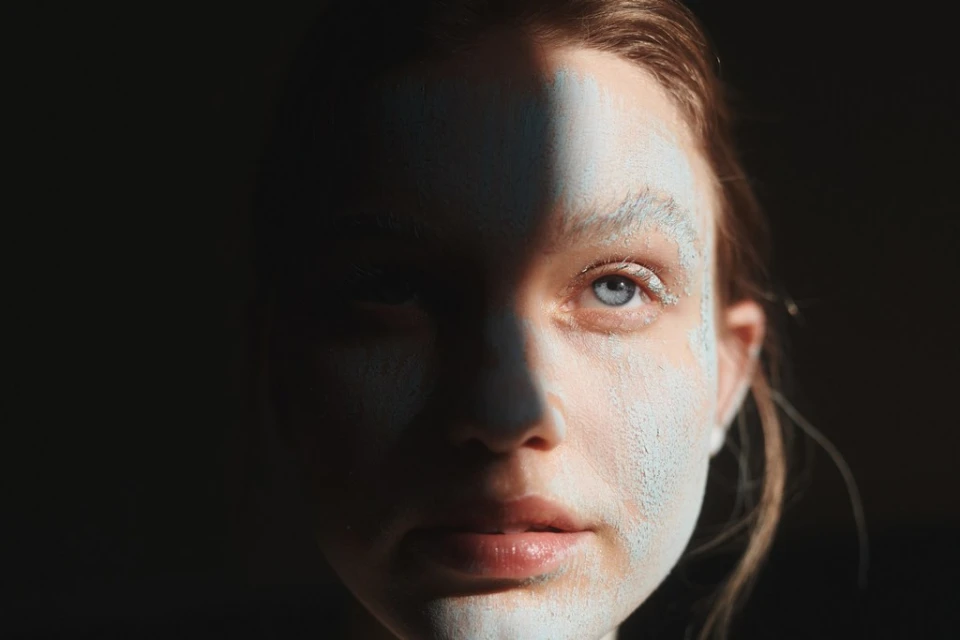 Close-up portrait of a woman with face mask partially applied, one side shadowed, highlighting her skin texture and eye color.