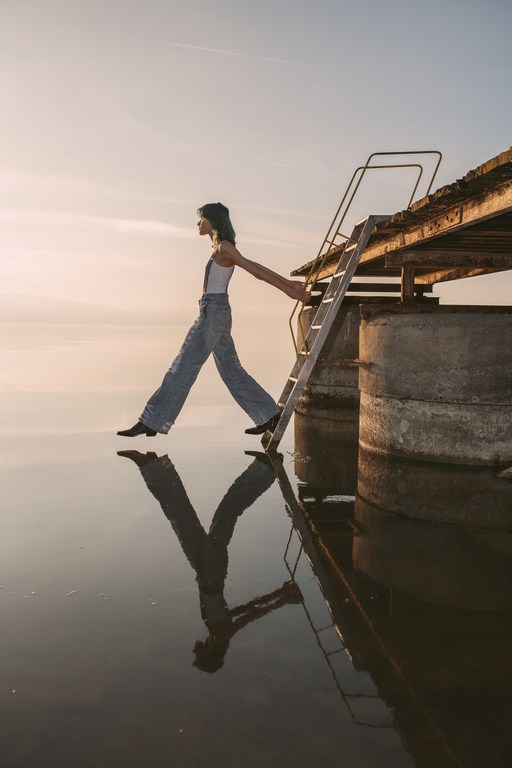 A full shot of a young adult female balancing on a concrete structure with a ladder leading into water, reflecting the sky at sunrise.