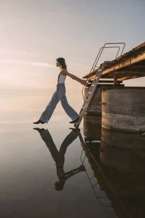 A full shot of a young adult female balancing on a concrete structure with a ladder leading into water, reflecting the sky at sunrise.