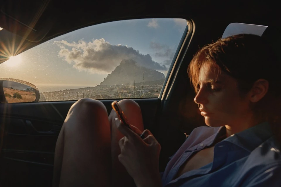 Young woman in a car using her cell phone, sun flares and landscape visible through the window. Vehicle interior, casual wear, relaxation.