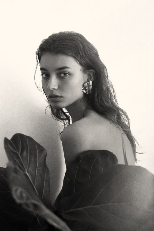 Black and white portrait of a young woman with wavy hair and large earrings, standing near a large leaf against a light background.