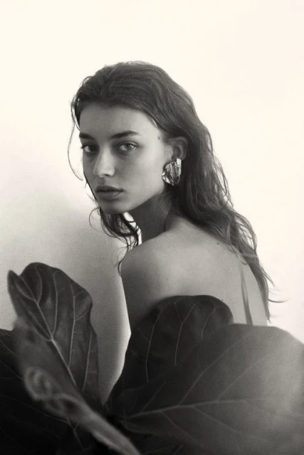 Black and white portrait of a young woman with wavy hair and large earrings, standing near a large leaf against a light background.