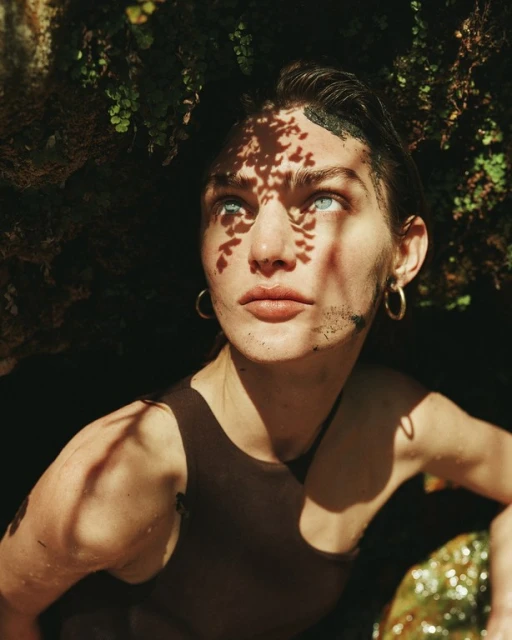 Portrait of a fair-skinned adult woman with light eyes, wearing a brown tank top, framed by foliage, with leaf shadows on her face.