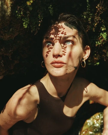 Portrait of a fair-skinned adult woman with light eyes, wearing a brown tank top, framed by foliage, with leaf shadows on her face.
