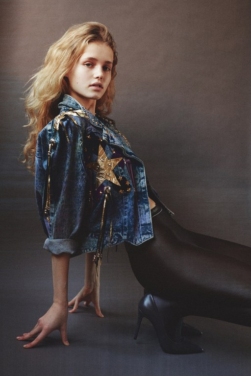 Young woman with long blonde hair poses in a stylish denim jacket and black leggings in a studio setting. Fashion photography.