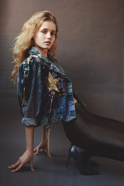 Young woman with long blonde hair poses in a stylish denim jacket and black leggings in a studio setting. Fashion photography.