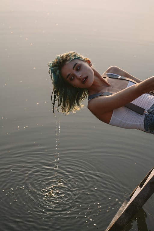 Young woman with blue hair leans over calm water on a sunny day, creating ripples as water drips from her hair. Outdoor scene.