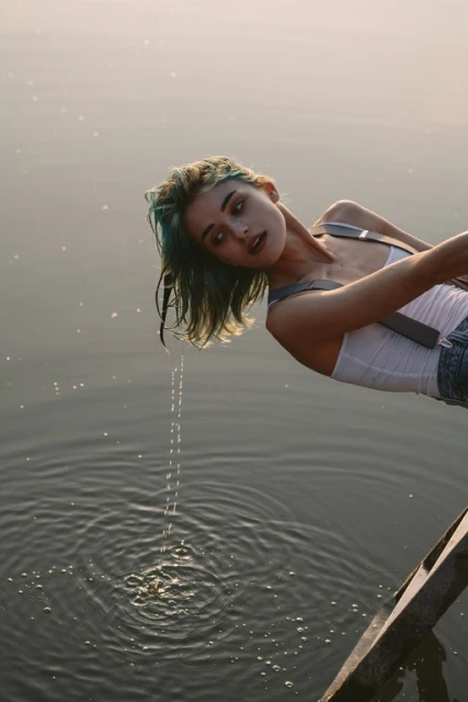 Young woman with blue hair leans over calm water on a sunny day, creating ripples as water drips from her hair. Outdoor scene.