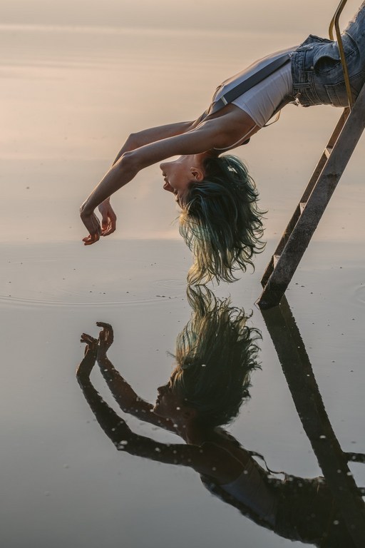 Teen girl bending over backwards from a wooden ladder towards her reflection in the water, with green dyed hair at sunset.