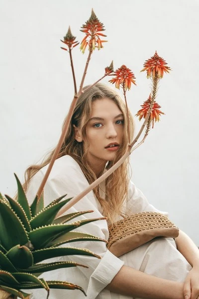 Blonde woman poses in white clothes with red aloe flowers, holding a woven bag in a natural outdoor setting with soft, neutral lighting.