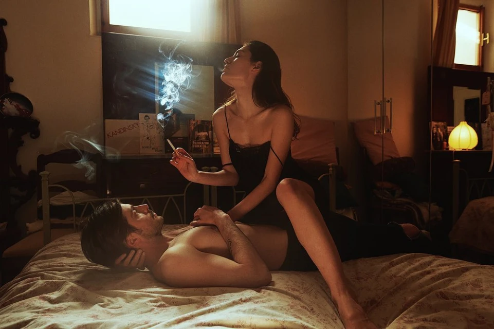 A couple in a bedroom setting; a woman is seated on top of a man, smoking a cigarette with smoke around them in warm, dim lighting.