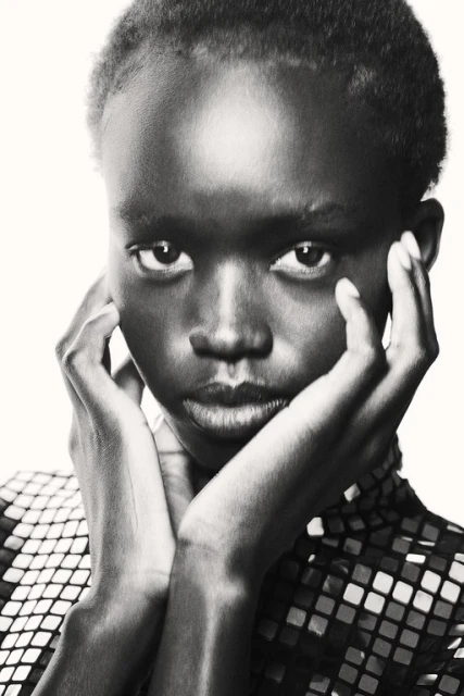 Close-up portrait of a young African-American woman with dark skin, short hair, and a patterned top, posing against a bright background.