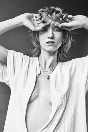 Monochrome portrait of a woman with tousled hair holding her hands to her forehead, wearing an open white t-shirt and a small cross necklace.