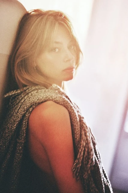 Woman with blond hair leans against a wall in soft light wearing a fringed shawl, looking pensively at the camera in medium shot.