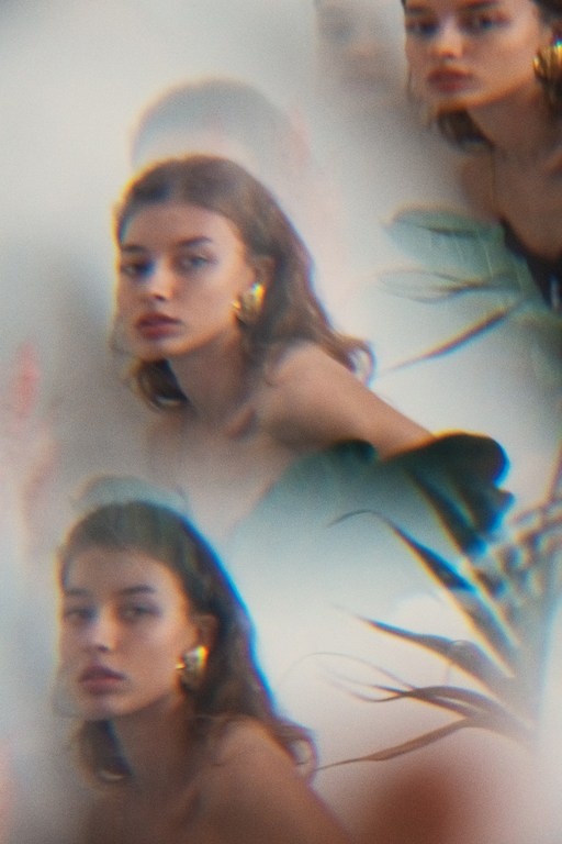 Dreamy portrait of a young woman with wavy brown hair and gold earrings, framed by a blurred tropical leaf, creating a surreal, artistic effect.