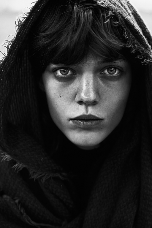 Monochrome portrait of a young adult with a dark hooded shawl, gazing intently with a serious expression, captured in a close-up shot.