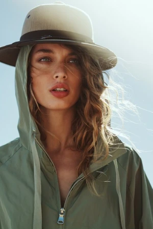 Close-up portrait of a woman wearing a green hooded jacket and a wide-brimmed hat against a bright sky background in Milan by Alessio Albi.