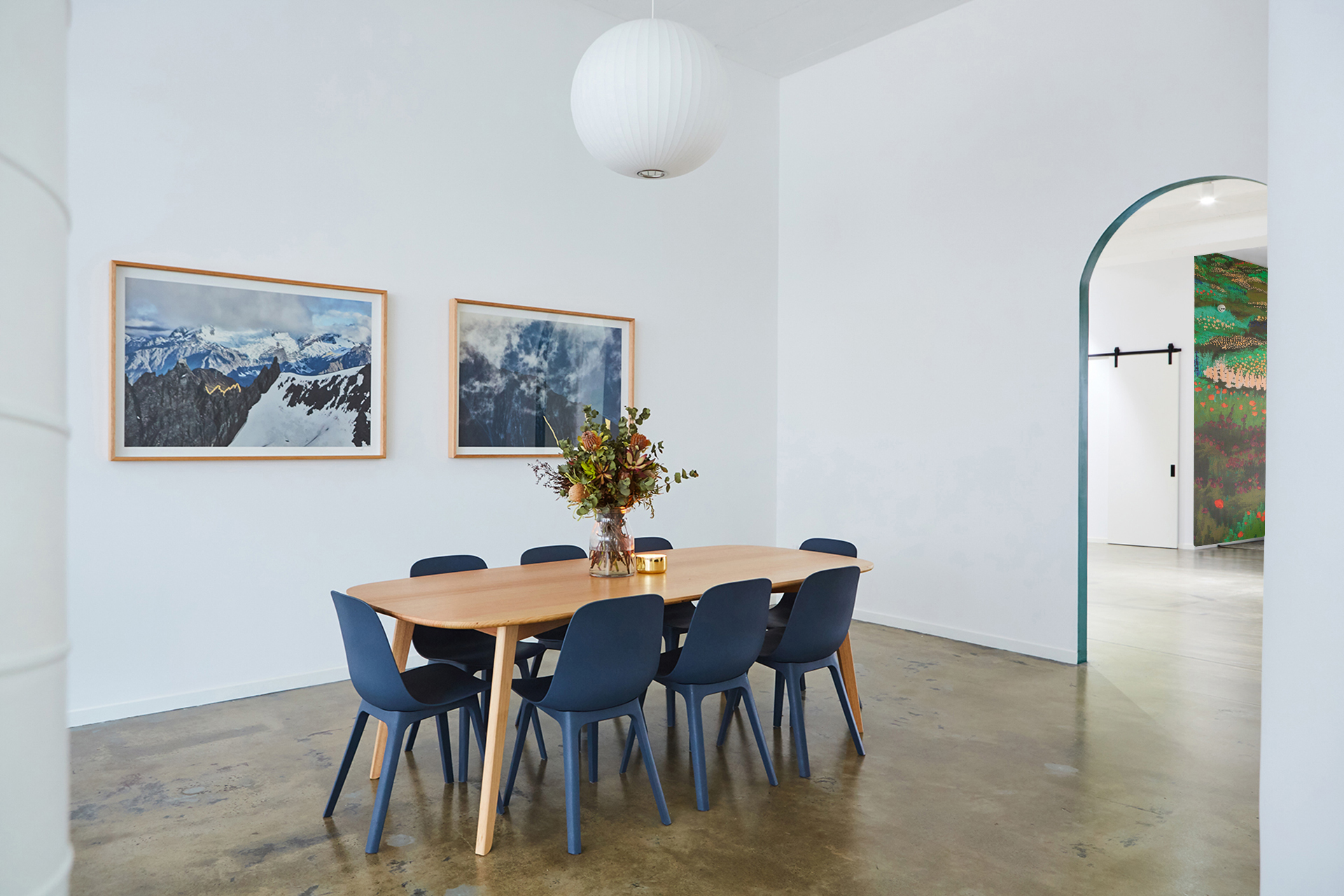 Brightly lit dining room featuring a wooden table surrounded by blue chairs, artwork on the wall, and a hanging light fixture.