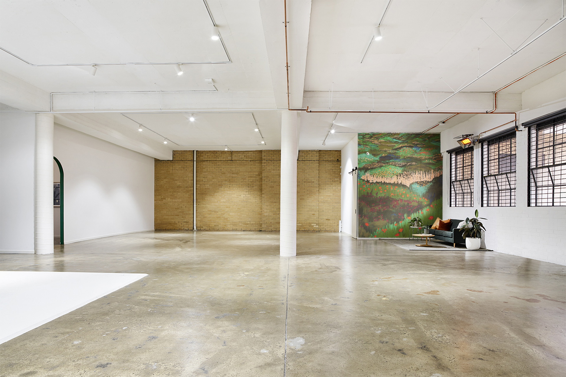 Wide shot of a bright photo studio featuring a concrete floor, white walls, large windows, and a decorative painting on the back wall.