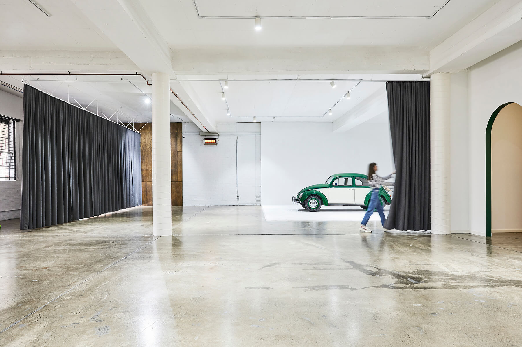 A woman parts gray curtains in a large, bright industrial studio with a vintage green and white Volkswagen Beetle car on display.