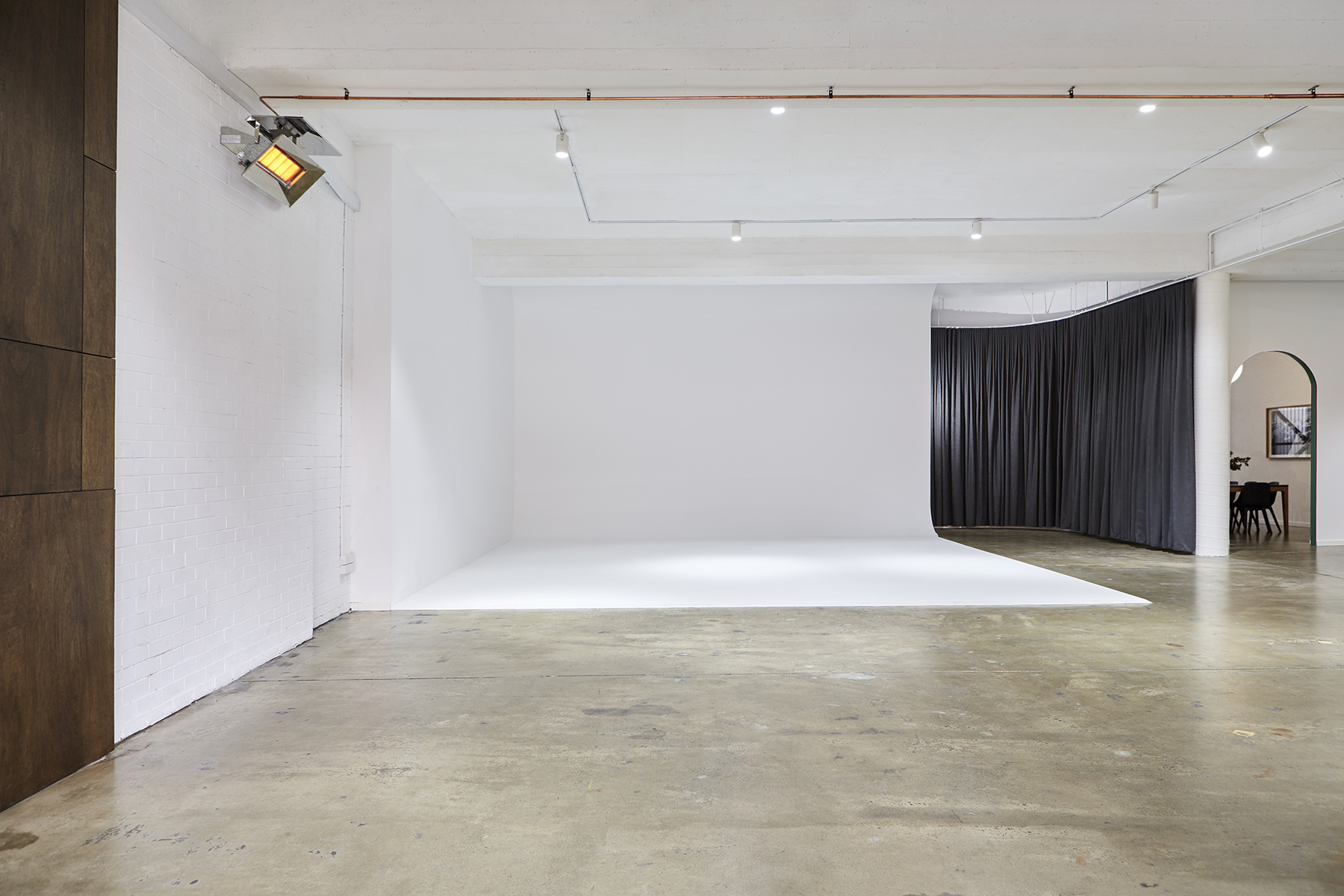 Photo studio with a white backdrop and grey concrete floor, spotlight on wall, black curtains in the background, indoor setting.