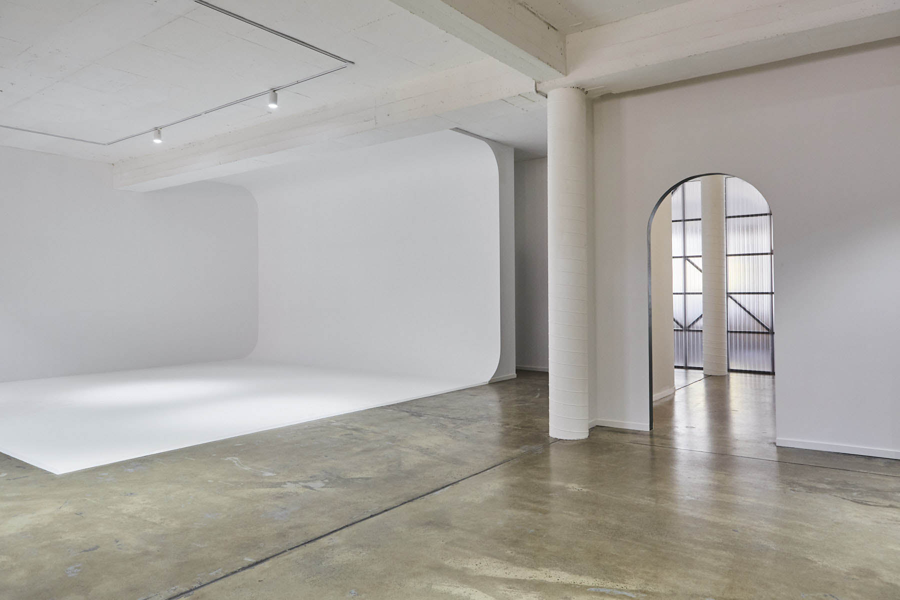 Bright, open photo studio interior with white walls, curved backdrop, concrete floor, pillars, archway, and grid-pattern windows in daylight.