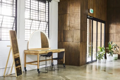 Modern wooden vanity table with illuminated arched mirror and two black stools inside a bright studio space in Melbourne