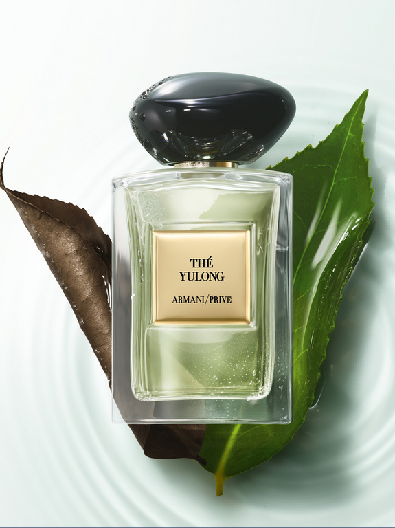 Elegant studio shot of Thé Yulong Armani Privé perfume bottle with black cap, surrounded by vibrant and dry tea leaves on light background.