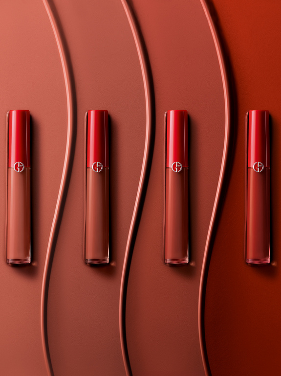 Overhead studio shot of four Giorgio Armani nude lip glosses with red caps, arranged in a row on a wavy, reddish-brown surface.
