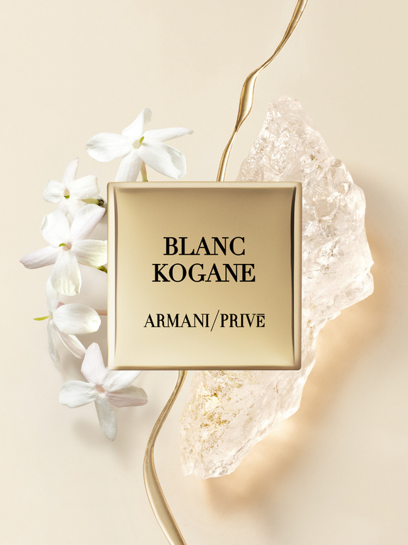 Armani Prive Blanc Kogane perfume bottle with white jasmine flowers, gold accents, and crystalline rock on a beige backdrop.