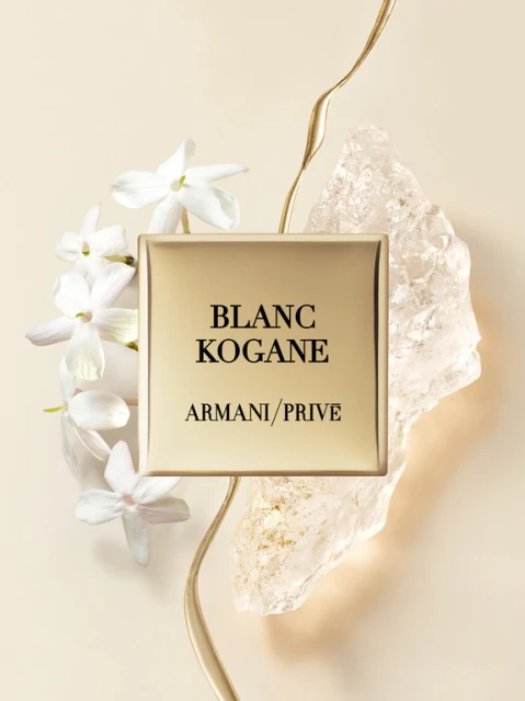 Armani Prive Blanc Kogane perfume bottle with white jasmine flowers, gold accents, and crystalline rock on a beige backdrop.