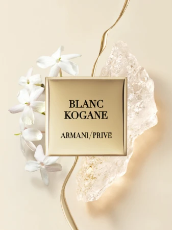 Armani Prive Blanc Kogane perfume bottle with white jasmine flowers, gold accents, and crystalline rock on a beige backdrop.