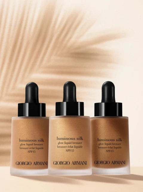 Three bottles of Giorgio Armani Luminous Silk glow liquid bronzer, arranged in a row, set against a soft background with palm leaf shadows.