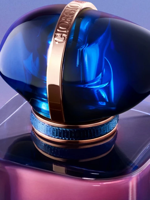 Close-up studio shot of a blue and gold perfume bottle with a transparent geometric base, set against a gradient purple backdrop.