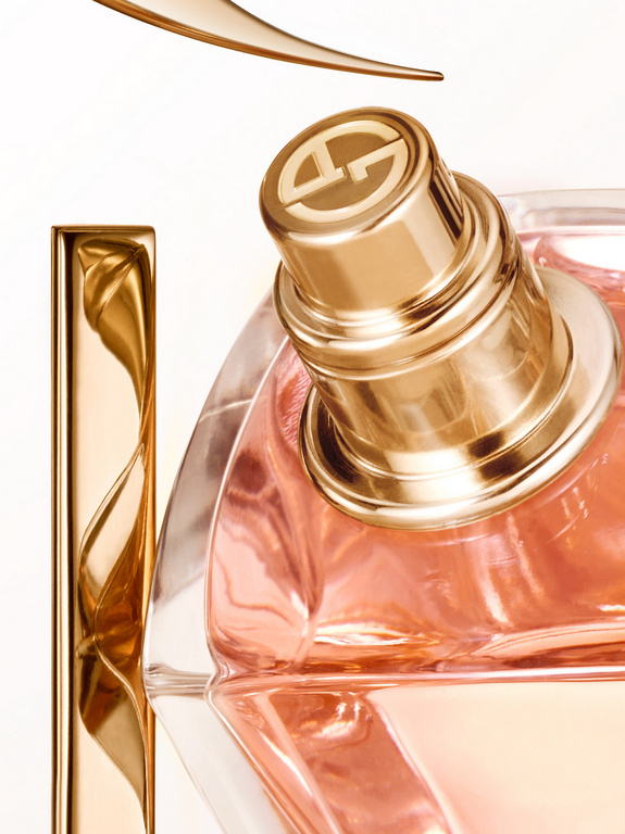 Close-up studio shot of a transparent perfume bottle with a golden cap, part of a golden design element above, on a white background.