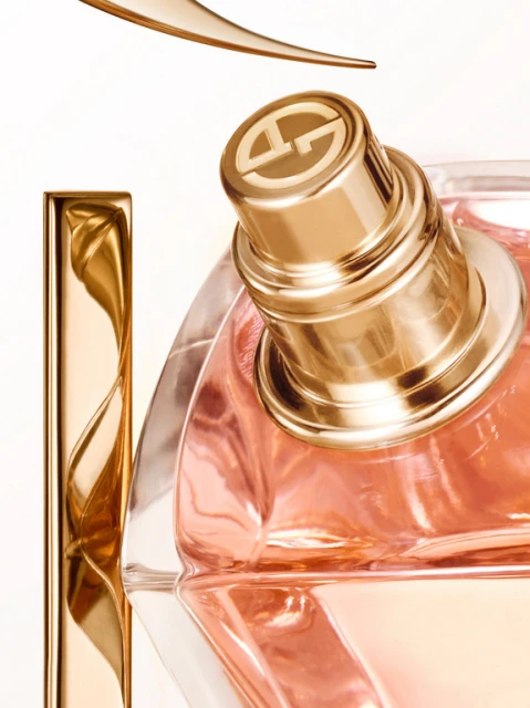 Close-up studio shot of a transparent perfume bottle with a golden cap, part of a golden design element above, on a white background.