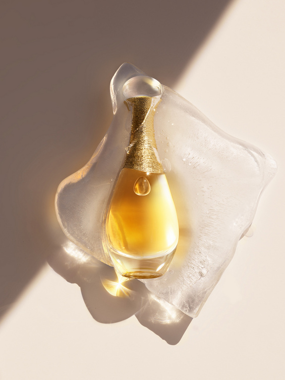 Elegant gold perfume bottle with curved glass resting on clear textured block with light and shadow play in a studio setting.
