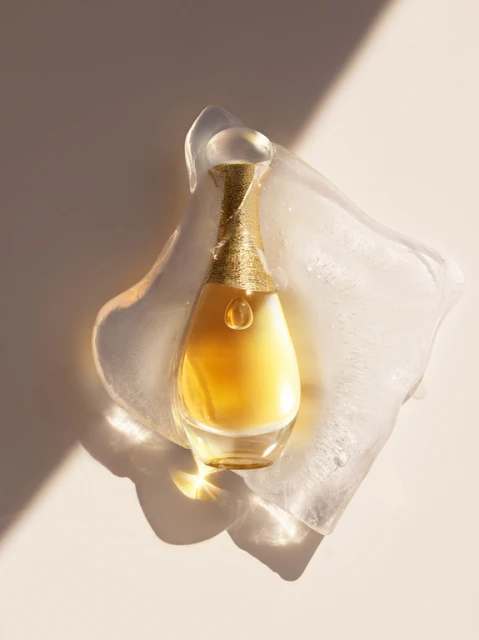 Elegant gold perfume bottle with curved glass resting on clear textured block with light and shadow play in a studio setting.