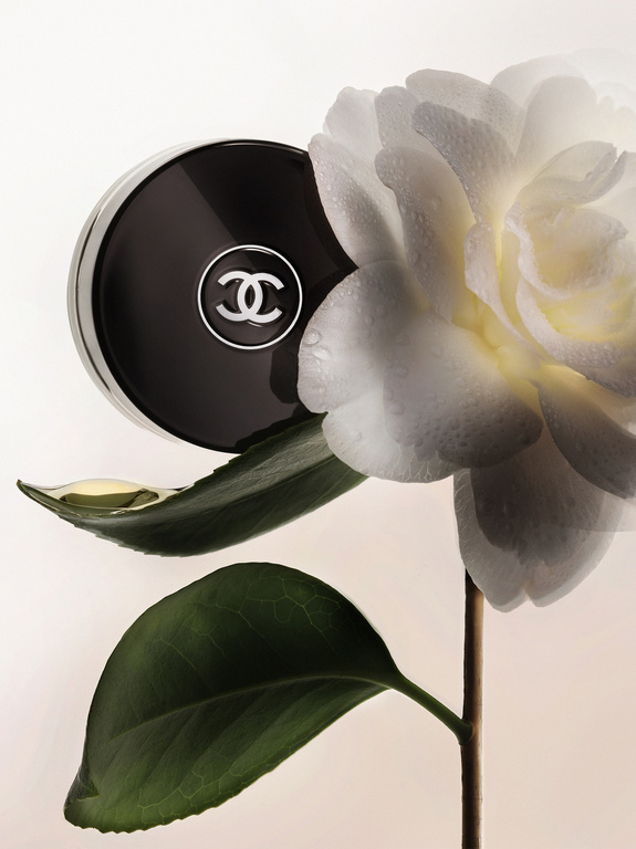 Close-up of a Chanel compact case with logo next to a white camellia flower with water droplets on a white background in studio shot.