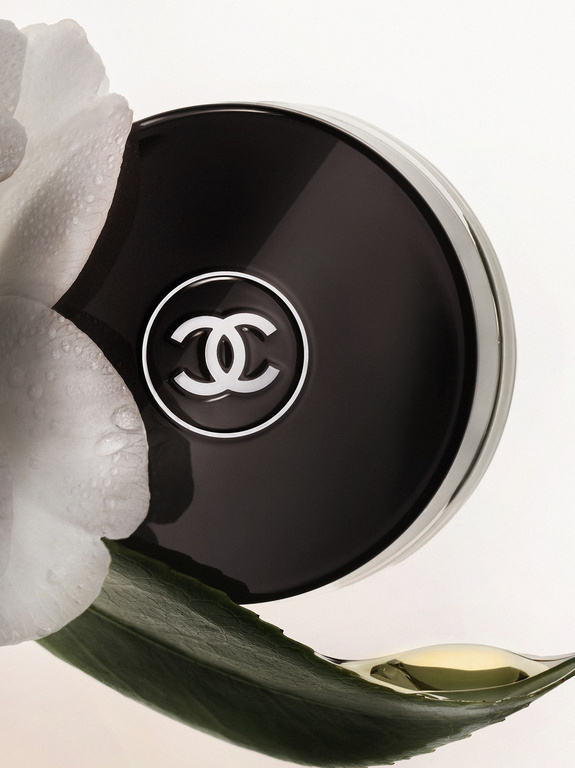 Close-up product shot of a Chanel compact with white flower petals and a dew-covered green leaf on a light background