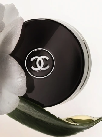 Close-up product shot of a Chanel compact with white flower petals and a dew-covered green leaf on a light background