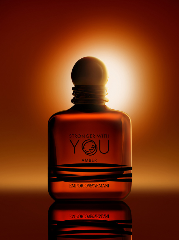 Amber-toned glass perfume bottle with black accents, lit dramatically against a gradient background in shades of orange and yellow.